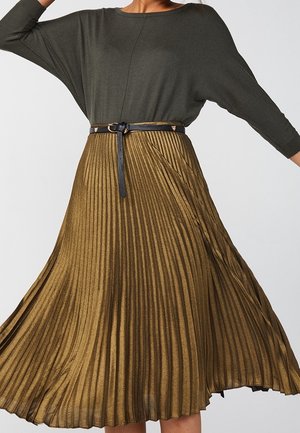 Vrouw die een olijfgroene top met lange mouwen draagt, ingestopt in een metallic bronzen geplooide midi-rok met zwarte riem, staand tegen een effen achtergrond.