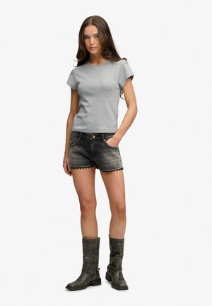 Vrouw met een grijs T-shirt met korte mouwen, zwarte denim shorts met gegolfde zoom en zwarte laarzen tot halverwege de kuit, staand met één hand in de zak.