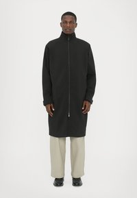 Black long coat with high collar and front zipper, featuring a relaxed fit and long sleeves, worn over light-colored trousers and black shoes.