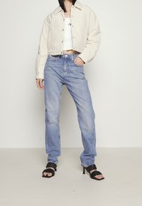 Beige kurze Jacke, weißes Oberteil, hellblaue Jeans mit geradem Bein und schwarze Sandalen mit Absatz und Rüschendetails. Einfaches und modernes Design.