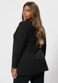 Svart ribbad långärmad topp med sidofickor, som har en avslappnad passform och en något rundad nederkant, ihop med svarta åtsittande byxor.