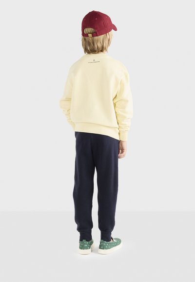 Gelbes Sweatshirt, marineblaue Jogginghose, grün gemusterte Schuhe und eine kastanienbraune Mütze. Das Sweatshirt hat ein kleines Logo auf der Rückseite.