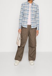 Rutigt skjorta i blått, vitt och grönt, parat med lösa olivgröna cargobyxor. Håller en liten beige handväska; vita sneakers är på fötterna.