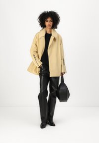 Beige oversized jakke med spredt krave, lynlåslommer og sorte læderbukser; model holder en rund sort taske; hvid baggrund.