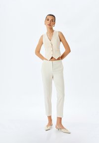 Cremefarvet skræddersyet vest med V-udskæring og fem knapper, parret med slim-fit bukser. Glat stof med minimal detaljering.