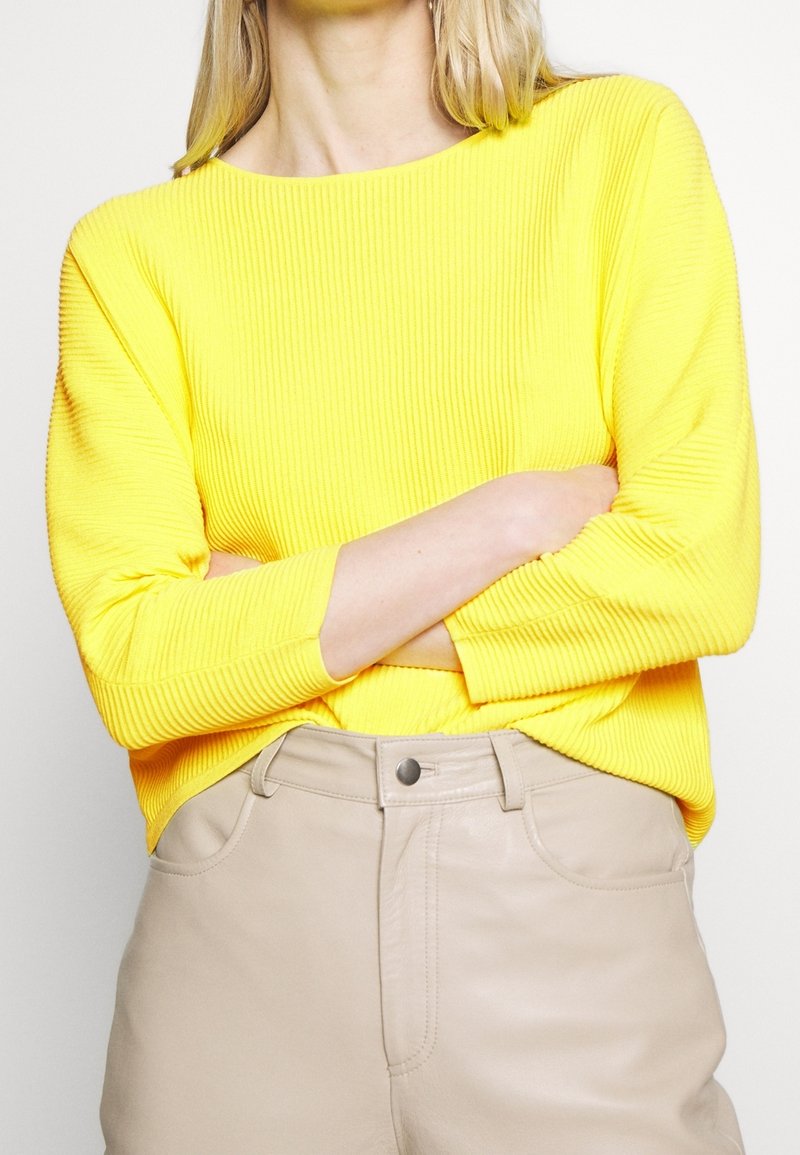 Personne portant un pull côtelé jaune vif à manches trois-quarts et un pantalon beige taille haute, les bras croisés sur la poitrine.