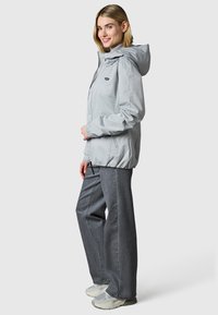 Vrouw met een lichtgrijze capuchonjas, losse donkergrijze spijkerbroek en grijze sneakers, staand en glimlachend tegen een effen achtergrond.