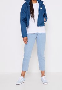 Personne portant une veste bleue North Face, un t-shirt blanc, un jean court bleu clair et des baskets Vans bleu clair, debout devant un fond blanc.