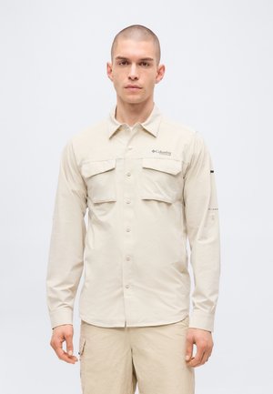Mand med kort hår iført beige skjorte med lange ærmer, klaplommer og matchende beige bukser, stående foran en ensfarvet baggrund.