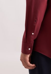 Bordeauxkleurig shirt met een gladde textuur, lange mouwen en knoopmanchetten. Heeft een subtiele geborduurde detail op de mouw.