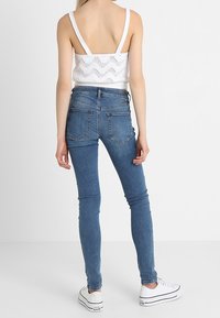 Top sin mangas de punto blanco con textura de patrón, combinado con jeans ajustados de denim azul y zapatillas blancas. Enfocado en la simplicidad y el ajuste.