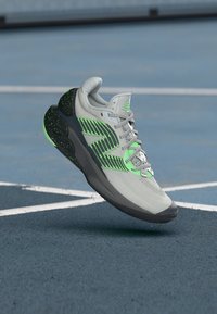 Grau-schwarzer Sportschuh mit grünen Akzenten und Schnürsenkeln, über einem blauen Outdoorplatz mit weißen Linien schwebend.