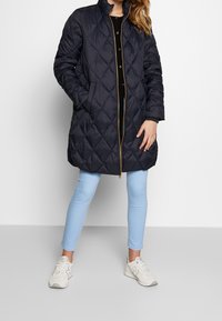 Marinblå quiltad lång kappa med hög krage, gulddragkedja och tryckknappar. Kombinerad med ljusblå leggings och vita sneakers.