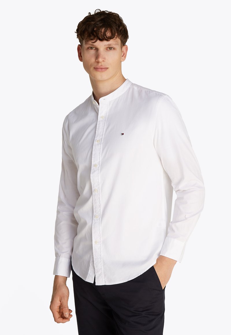 Jeune homme aux cheveux bouclés portant une chemise blanche à col mandarin boutonnée et un pantalon noir, debout avec une main dans la poche.