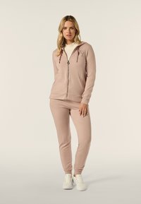 Felpa rosa chiaro con zip e cordino, abbinata a pantaloni tapered coordinati. Realizzata in tessuto morbido, presenta un discreto dettaglio del logo.