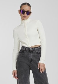 White ribbed zip-up crop top with high collar, paired with high-waisted, faded black jeans. Model wears oversized purple sunglasses.