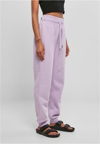 Pantalons de survêtement lavande avec taille élastique et cordon de serrage, comprenant des poches latérales et une coupe décontractée, associés à des sandales noires à glisser.