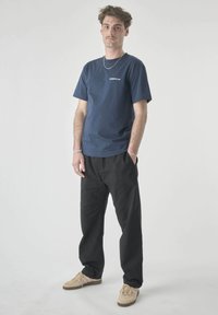 Young man with short hair and mustache wearing navy blue graphic t-shirt, black pants, beige sneakers, and silver jewelry standing against gray background.