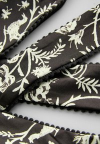 Čierny textil s bielymi kvetinovými a zvieracími vzormi, s textúrovaným strapcovým lemom pozdĺž okrajov, prezentujúci zložitý dizajn.