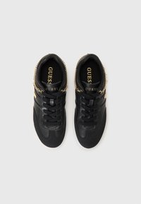 Svarte joggesko med en glatt overdel i lær, rund tå, flat hvit såle, snøring foran og gullkjede detaljer på sidene. Merkevarenlogo synlig inni.