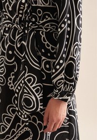 Schwarzes Kleid mit weißem abstraktem Muster, mit langen Ärmeln, einem Taillengürtel und einem knöpfbaren Manschette, das einen strukturierten Stoff präsentiert.