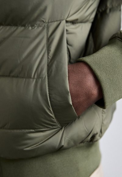 Olivenfarvet pufferjakke med quiltet design. Har en tekstureret grøn manchet og en synlig hånd i en sidelomme. Glat, blankt stof.