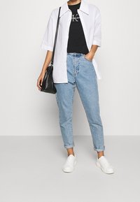 Camisa blanca de botones sobre una camiseta gráfica negra, jeans de tiro alto azules y zapatillas blancas. Un bolso negro de hombro añade contraste.