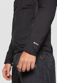 Giacca nera isolata con una vestibilità aderente, caratterizzata da una texture liscia e il marchio Pertex Quantum sul polsino. Tasca con cerniera visibile.