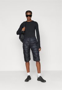 Pessoa vestindo uma blusa de manga longa preta, calções acolchoados até ao joelho, meias brancas, ténis pretos, grandes óculos de sol pretos, carregando uma mochila preta ao ombro.