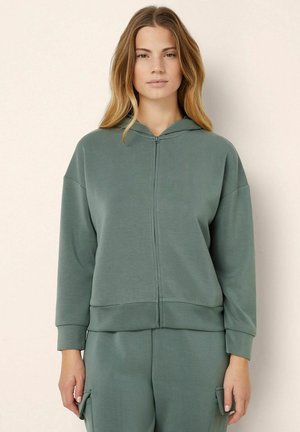 Felpa con zip in tessuto verde, con una vestibilità comoda, cappuccio con cordino e polsini a coste. Texture liscia senza motivi visibili.