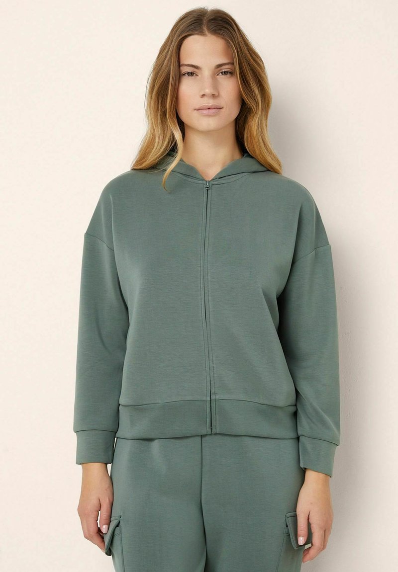 Felpa con zip in tessuto verde, con una vestibilità comoda, cappuccio con cordino e polsini a coste. Texture liscia senza motivi visibili.