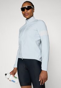 Light blue cycling jacket with zipper, stand collar, and reflective accents. Worn over black cycling shorts. Hand holds a water bottle.