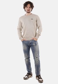 Uomo con capelli ricci, che indossa un maglione beige, jeans blu strappati e sneaker marroni e bianche, in piedi con le mani in tasca.