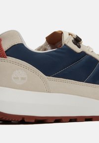 Sneaker med övre i marinblått tyg, beige mockadetaljer, röd dragflik, gummisula och logotypprägel på sidan.