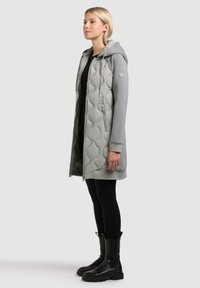 Blondynka stojąca w szarej długiej pikowanej kurtce z kapturem, czarnych legginsach i czarnych botkach za kostkę na jasnoszarym tle.