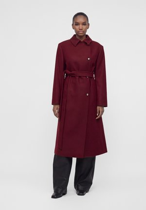 Femme debout portant un long manteau bordeaux ceinturé avec des boutons argentés et un pantalon noir large sur un fond gris clair uni.