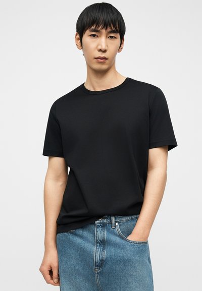 Νεαρός άνδρας με ίσια μαύρα μαλλιά, φορώντας ένα απλό μαύρο κοντομάνικο μπλουζάκι και μπλε τζιν, με το ένα χέρι στην τσέπη, ουδέτερο φόντο.