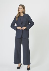 Sacou bleumarin cu umeri structurați, asortat cu pantaloni largi cu dungi în culori închise, completat cu pantofi cu toc înalt negri.