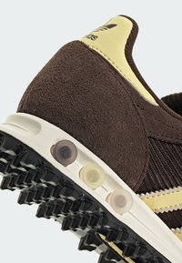 Καφέ πάνινα παπούτσια σουέντ και δίχτυ με κίτρινες πινελιές, υφασμένους επιφανειακούς χώρους και λαστιχένια σόλα με κυκλικά καρφιά για πρόσφυση.