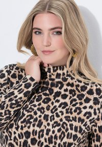 Bluzka w panterkę z długimi rękawami, wysokim kołnierzem i gładkim materiałem w odcieniach beżu i ciemnego brązu. Złote okrągłe kolczyki dopełniają stylizację.