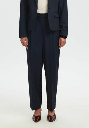 Personne portant un pantalon et une veste de costume bleu marine foncé, un haut blanc, un bracelet de cheville foncé et des chaussures pointues marron, debout sur un fond uni.