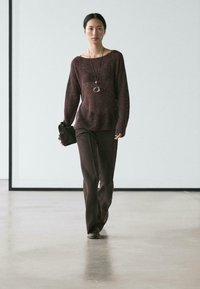 Bordeaux geribbelde trui met brede halslijn, gecombineerd met een bruine broek. Model houdt een zwarte clutch met zilverkleurige hardware vast. Glad, mat oppervlak.