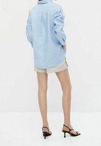 Himmelblaues Langarmshirt mit einem Rückenfalten, getragen mit beigen Shorts. Schwarze High Heels runden das Outfit ab. Schlichte und neutrale Farbpalette.