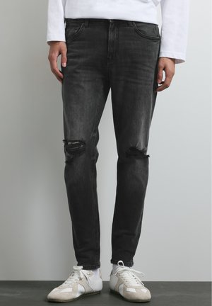 Personne portant un jean noir déchiré et des baskets blanches, debout contre un mur et un sol gris clair.
