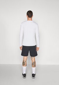 Camisa blanca de manga larga combinada con pantalones cortos negros, con una textura suave y un ligero patrón gráfico. Zapatillas negras y calcetines blancos completan el conjunto.
