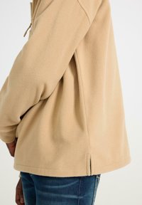Beige fleece takki, jossa on teksturoitu pinta, sivuaukko helmassa ja rento istuvuus. Yhdistettynä sinisiin farkkuihin.
