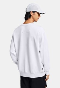 Sweatshirt blanc au fit décontracté, avec des poignets côtelés et un petit logo en détail sur l'ourlet. Porté avec une casquette gris clair avec des accents perforés.