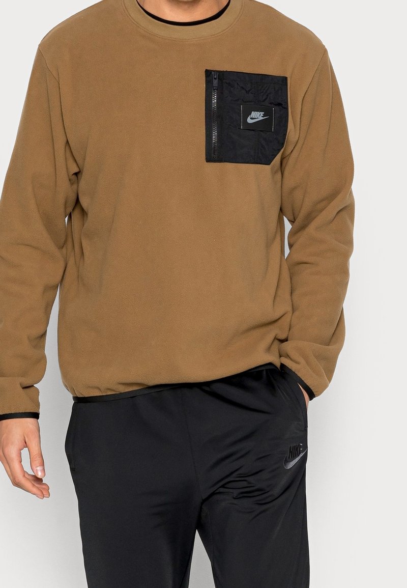 Hnědá fleecová mikina s černou kapsou na zip na hrudi, která má malé logo. Černé kalhoty s logem na boku.