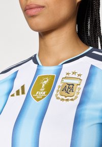 Camisa da seleção argentina com listras verticais azuis e brancas, um logo da Adidas e o emblema dourado de Campeões do Mundo 2022 da FIFA.
