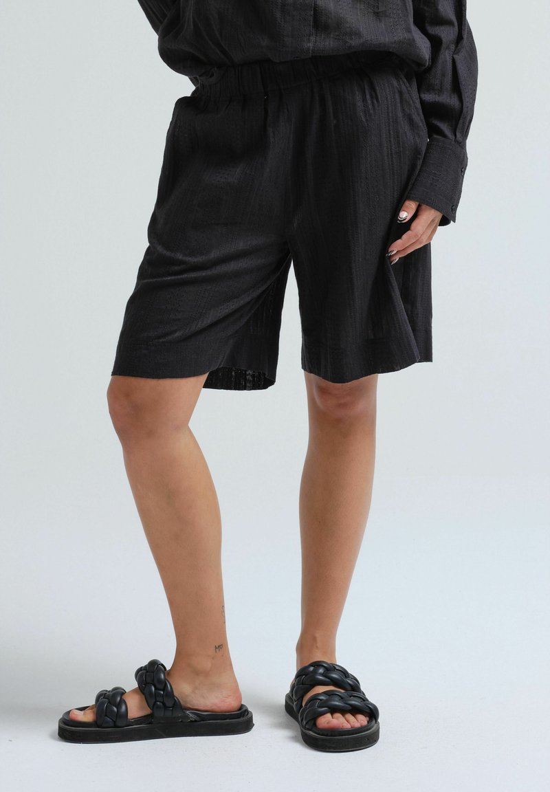Pantaloni corti neri realizzati in tessuto leggero con un motivo testurizzato, dotati di una vita elastica. Abbinati a sandali neri a ciabatta spessi.